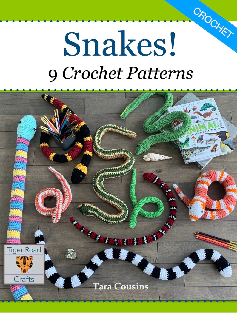 May include: Nine crochet snake patterns, including a black and white striped snake, a red and black snake, a green snake, a yellow and black snake, a pink and yellow snake, and a coral snake. The patterns are displayed on a wooden surface with a book about animals in the background.