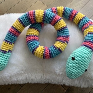 May include: A crocheted snake toy with a light blue, yellow, pink, and purple striped body. The snake is curled into a circle with its head facing the viewer. The snake has black eyes.