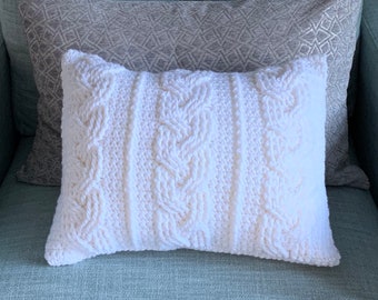 May include: A rectangular white cable-knit pillow with a textured design, resting on a light blue surface. A gray patterned pillow is behind it. The pillow is a decorative home decor item.