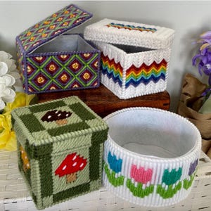 May include: A collection of handmade, needlepoint boxes in various shapes and sizes. One box features a geometric pattern in purple, green, and yellow. Another has a rainbow zig-zag design. A third box is decorated with mushrooms, and a round container has a floral pattern.