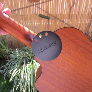 May include: A close-up of a ukulele with a dark brown wooden body and neck. A black circular attachment with the text "UkeLoop" is affixed to the body. The ukulele is held by a person.