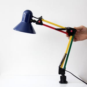 May include: A multi-coloured desk lamp with a navy blue lampshade. The arm is made up of yellow, red, and green sections. The lamp is on a white surface.