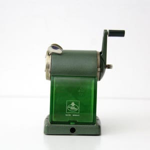 Pu&ograve; includere: Un temperamatite manuale verde scuro con manovella e un contenitore per i trucioli in plastica verde traslucida. La parte anteriore presenta il logo Faber-Castell, il numero di modello 52/25 e la parola GERMANY stampata in bianco.