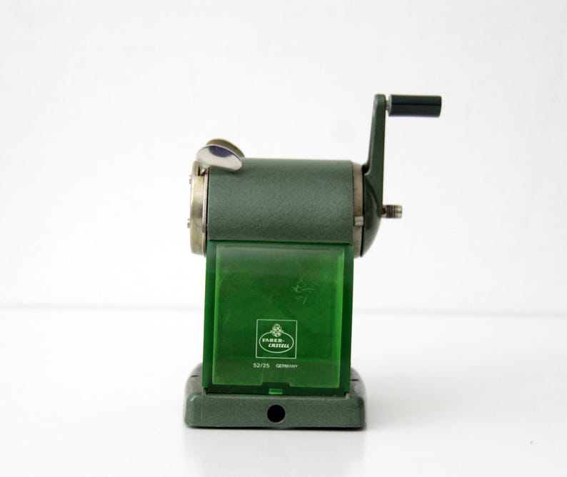 Pu&ograve; includere: Un temperamatite manuale verde scuro con manovella e un contenitore per i trucioli in plastica verde traslucida. La parte anteriore presenta il logo Faber-Castell, il numero di modello 52/25 e la parola GERMANY stampata in bianco.