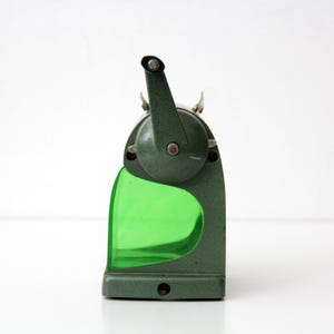 Pu&ograve; includere: Un temperamatite manuale vintage con corpo in metallo verde scuro testurizzato e un contenitore per i trucioli in plastica verde brillante traslucida. Il dispositivo presenta una manovella montata superiormente e una sagoma curva.