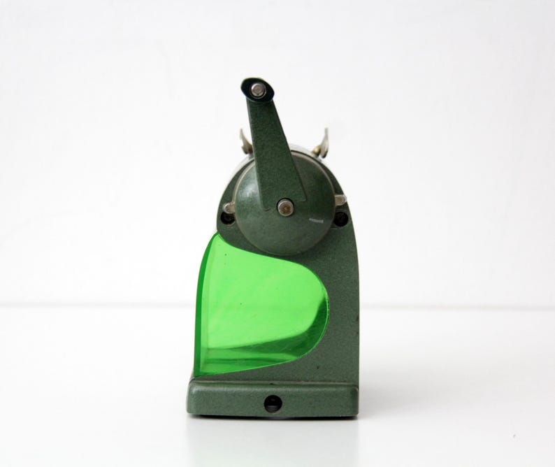 Pu&ograve; includere: Un temperamatite manuale vintage con corpo in metallo verde scuro testurizzato e un contenitore per i trucioli in plastica verde brillante traslucida. Il dispositivo presenta una manovella montata superiormente e una sagoma curva.