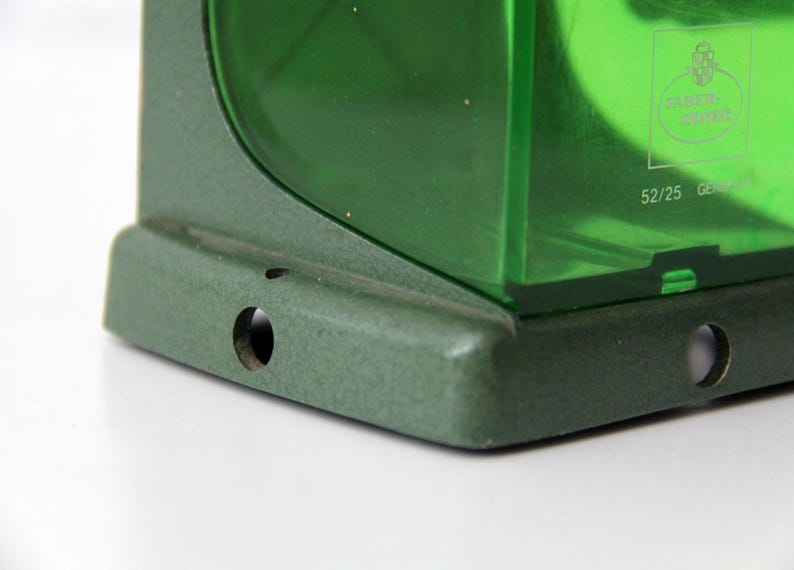 Pu&ograve; includere: Una base in metallo verde con due fori di montaggio circolari sostiene un componente in plastica verde traslucida. La sezione in plastica presenta un logo stampato e il testo 52/25 GERMANY, con una finitura testurizzata sul telaio in metallo.