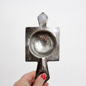 Op de afbeelding: Een zilverkleurige metalen theezeef met een vierkant frame, een centrale ronde geperforeerde kom en twee puntige handvatten aan de boven- en onderkant. Het oppervlak heeft een verweerde, doffe afwerking met een zichtbare metalen textuur.