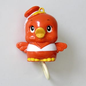May include: Orange plastic bird-shaped toy with a yellow beak and feet, white chest band, and a matching orange hat. The toy has black eyes and small wings. A white pull string hangs from the bottom.