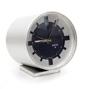 May include: A cylindrical silver-toned metal alarm clock with a dark blue circular face. The clock features a silver bezel with black rectangular hour markers, silver hands, and the text Swiza 8 printed in white on the face. It rests on a base.