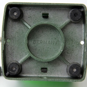 Pu&ograve; includere: La parte inferiore di una base quadrata per elettrodomestici da cucina di colore verde, dotata di quattro piedini circolari in gomma nera agli angoli, un anello di supporto centrale circolare e la scritta GERMANY impressa al centro del telaio in metallo verde testurizzato.