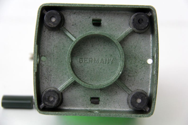 Pu&ograve; includere: La parte inferiore di una base quadrata per elettrodomestici da cucina di colore verde, dotata di quattro piedini circolari in gomma nera agli angoli, un anello di supporto centrale circolare e la scritta GERMANY impressa al centro del telaio in metallo verde testurizzato.