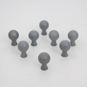 May include: Eight grey cabinet knobs on a white surface. Each knob features a rounded top and a short, cylindrical base. The knobs are evenly spaced, forming a symmetrical pattern. The knobs are made of a solid, matte material.