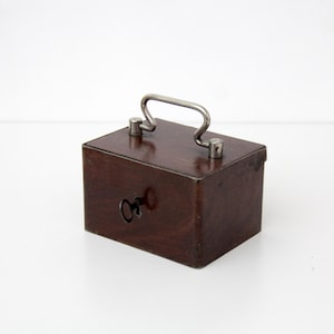 May include: A small, rectangular wooden box with a dark brown finish. It features a silver-colored handle and a keyhole-shaped cutout on the front. The box is designed with a simple, classic aesthetic.
