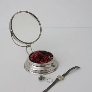 May include: A silver-toned circular vanity box with an attached upright mirror, containing red textured material. Beside the box sits a silver ring with a dark green oval stone, a plain silver band, and a silver-toned wristwatch with a mesh strap.