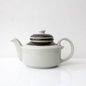 May include: A ceramic teapot featuring a light gray body and a dark brown glazed band around the upper shoulder and lid. The teapot has a curved handle, a short spout, and a matching lid with a small knob handle on top, presented in a simple design.