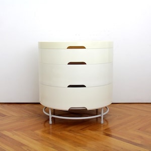 May include: A cylindrical off-white storage unit featuring four stacked drawers with integrated semi-circular handle cutouts. The unit is supported by a circular white metal frame base with four short legs, providing a stable foundation for the structure.