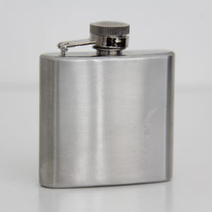 Puede incluir: Una petaca rectangular de acero inoxidable con acabado de metal cepillado y tapón de rosca articulado. La petaca presenta un gráfico sutil y tenue grabado con láser en la superficie frontal, con un diseño elegante, metálico y funcional.