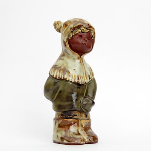 May include: A ceramic figurine of a person in winter attire. The figure has a dark brown face and is wearing a hooded coat in shades of green, brown, and cream. The boots are a darker brown with a textured appearance.