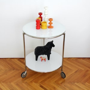 May include: A two-tiered round rolling cart with a white surface and silver metal frame. The top shelf holds five colorful wooden candle holders in red, yellow, and white. The bottom shelf features a black textured sheep figure and a small red horse.