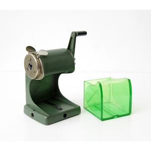 Pu&ograve; includere: Un temperamatite manuale verde oliva scuro con manovella in metallo e una piastra frontale circolare color argento. Accanto al temperamatite si trova un contenitore per trucioli rimovibile in plastica verde brillante traslucida a base quadrata.