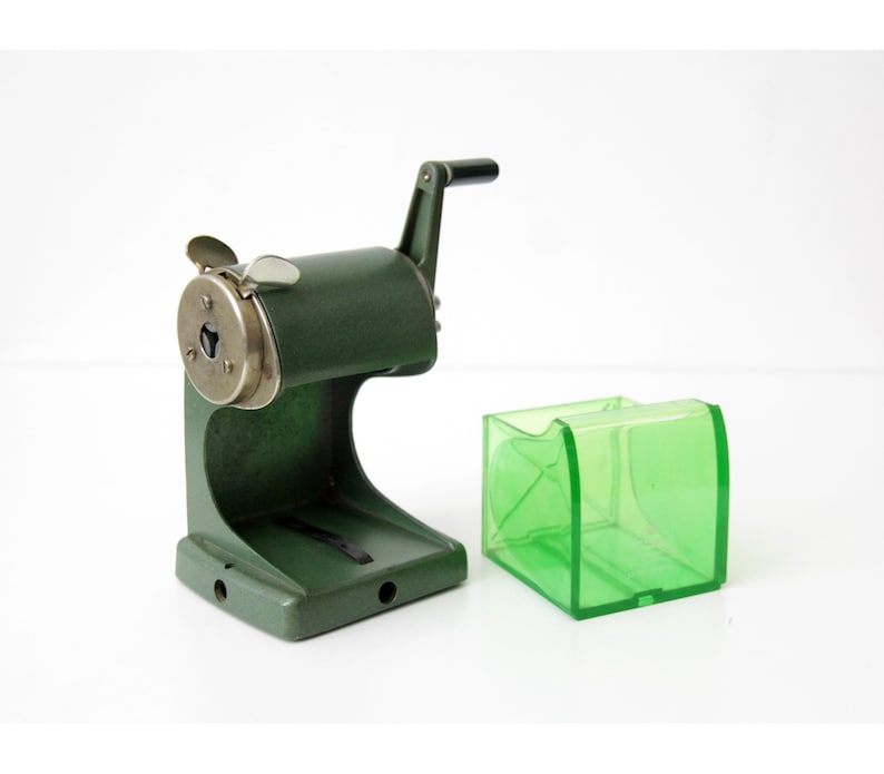 Pu&ograve; includere: Un temperamatite manuale verde oliva scuro con manovella in metallo e una piastra frontale circolare color argento. Accanto al temperamatite si trova un contenitore per trucioli rimovibile in plastica verde brillante traslucida a base quadrata.