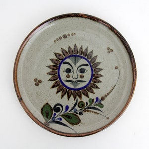 Puede incluir: Un plato redondo de cerámica con un diseño de sol pintado a mano. El sol tiene una cara con bigote y está rodeado de rayos marrones. El plato tiene un fondo gris moteado, un borde marrón y detalles florales decorativos.