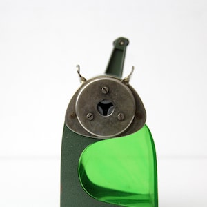 Pu&ograve; includere: Un temperamatite vintage con corpo in metallo verde oliva scuro e serbatoio in plastica verde brillante traslucido. La piastra frontale &egrave; circolare e metallica, fissata con tre viti, e include un'apertura centrale per le matite.