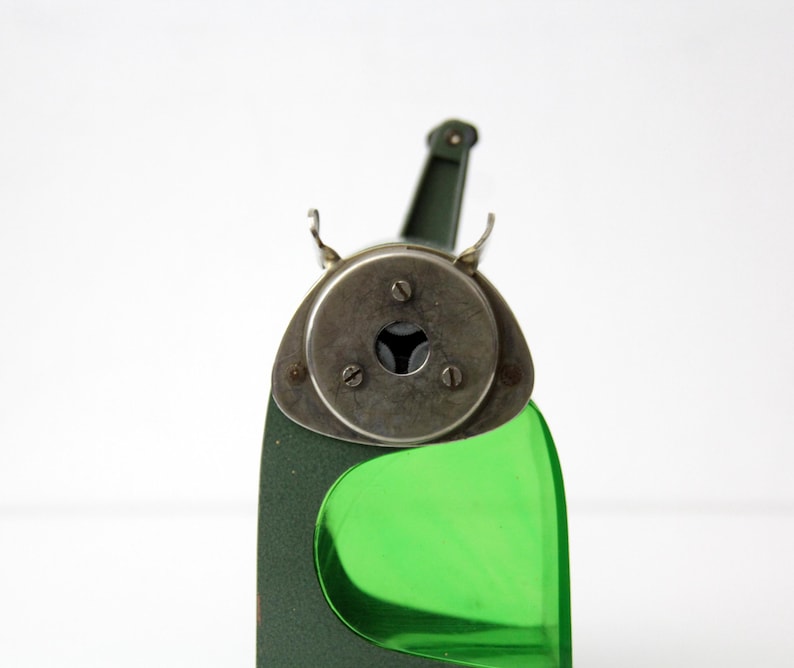 Pu&ograve; includere: Un temperamatite vintage con corpo in metallo verde oliva scuro e serbatoio in plastica verde brillante traslucido. La piastra frontale &egrave; circolare e metallica, fissata con tre viti, e include un'apertura centrale per le matite.