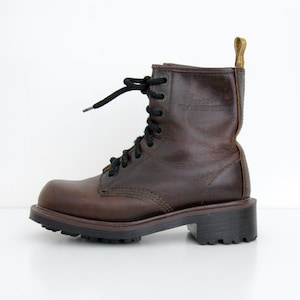 Puede incluir: Bota de combate de cuero marrón con cordones negros y suela negra gruesa. La bota tiene un logotipo de Dr. Martens y una lengüeta amarilla. Un estilo clásico, adecuado para varios conjuntos.