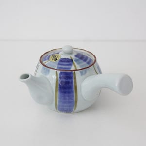 Peut inclure: Une théière en céramique blanche avec une anse latérale et un couvercle, ornée de rayures verticales bleues peintes à la main et de fines lignes vert olive. Le bord du couvercle est souligné d'une fine ligne brune et un petit autocollant doré est visible.