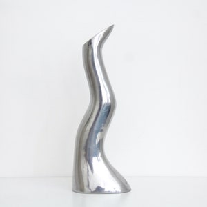 May include: A polished, silver sculpture with a curved, organic form. The sculpture has a smooth, reflective surface and a slender, elongated shape that tapers towards the top. It stands on a flat base.