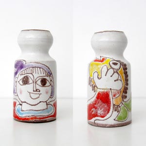 May include: Two white ceramic vases with a narrow neck and a rounded rim. The left vase features a hand-drawn illustration of a person with purple hair. The right vase displays a colorful abstract figure with yellow, red, and green painted details.
