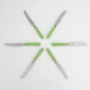 May include: Six stainless steel table knives arranged in a circular star pattern. Each knife features a smooth, light green handle and a curved metal blade with a visible bolster. The blades are pointed toward the center of the arrangement.