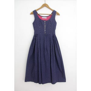 May include: A sleeveless navy blue dress featuring a white polka dot pattern, a round neckline with a pink interior lining, and a front button closure with six small buttons. The dress has a fitted bodice and a pleated, knee-length skirt.