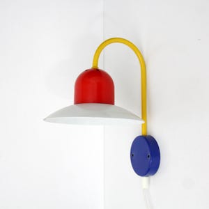 Può includere: Una lampada da parete con una tavolozza di colori primari. L'apparecchio presenta una base circolare blu, un braccio giallo curvo, un alloggiamento rosso a forma di cupola e un ampio paralume conico bianco. Il design usa colori solidi e vivaci.