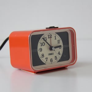 Puede incluir: Un despertador naranja vintage con una esfera negra y números blancos. El reloj tiene forma rectangular y un cable de alimentación negro. La esfera muestra la hora.