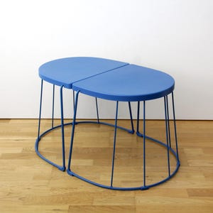 Könnte beinhalten: Ein Set aus zwei blauen Satztischen mit halbkreisförmigen Tischplatten und dünnen, vertikalen Drahtgestellbeinen, die eine ovale Basis bilden. Die Tische sind aus Metall mit einer glatten blauen Oberfläche gefertigt und lassen sich ineinanderschieben.