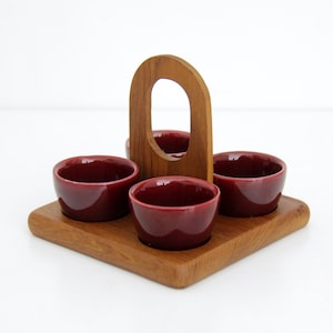 Può includere: Un vassoio di legno con quattro piccole ciotole in ceramica bordeaux. Il vassoio ha una maniglia integrata ed è realizzato in legno chiaro. Le ciotole sono uniformemente distanziate sul vassoio.