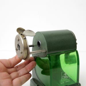 Pu&ograve; includere: Un temperamatite manuale vintage con corpo in metallo verde scuro e un contenitore per i trucioli in plastica verde traslucida. Il dispositivo presenta una piastra frontale in metallo argentato con manovella e un'apertura circolare.