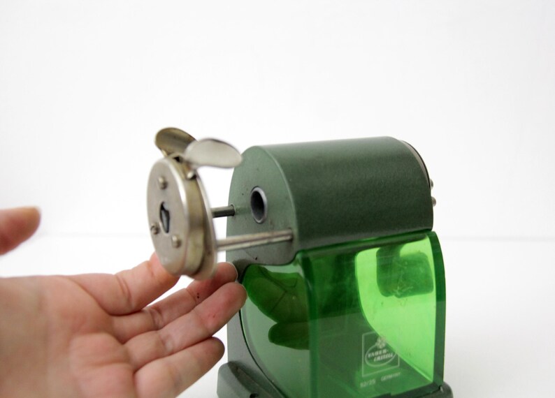 Pu&ograve; includere: Un temperamatite manuale vintage con corpo in metallo verde scuro e un contenitore per i trucioli in plastica verde traslucida. Il dispositivo presenta una piastra frontale in metallo argentato con manovella e un'apertura circolare.