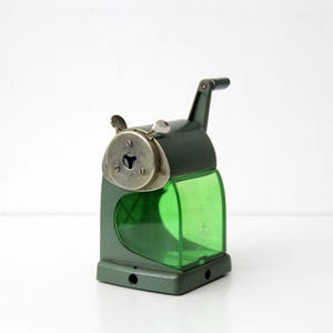 Pu&ograve; includere: Un temperamatite manuale con corpo in metallo verde scuro e un contenitore per i trucioli in plastica verde traslucida. &Egrave; dotato di una piastra frontale in metallo argentato con apertura circolare, due piccole viti a farfalla e una manovella nera.