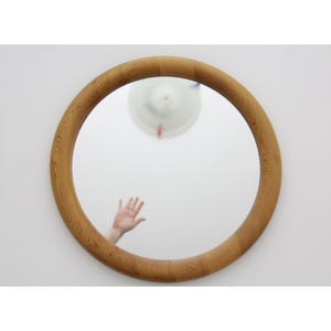 Puede incluir: Un espejo redondo con un marco de madera marrón claro. El espejo refleja un techo blanco y la mano de una persona. El marco tiene un patrón de vetas de madera natural, añadiendo un toque de encanto rústico al diseño.