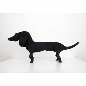 May include: A black wooden dachshund dog sculpture. The dog has a long body, short legs, and a curved tail. The sculpture is made of several pieces of wood joined together with visible joints. The dog is standing on a white surface.