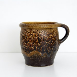 Può includere: Una tazza in ceramica con corpo arrotondato e un manico singolo. L'esterno presenta uno smalto testurizzato con una sfumatura di marrone scuro alla base che passa a una finitura maculata marrone dorato verso il bordo spesso e arrotondato.