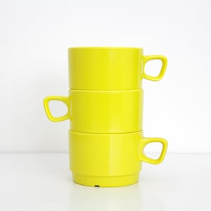 May include: Three bright yellow ceramic mugs stacked. Each mug has a square handle and a cylindrical body. The mugs are stacked on a white surface against a white background.