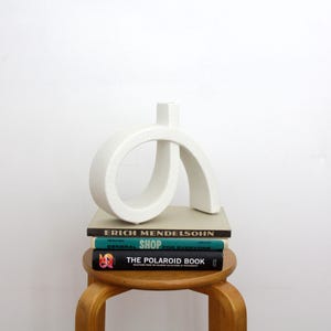 Op de afbeelding: Een witte keramische vaas met een gebogen, abstracte lusvorm en een getextureerd oppervlak. De vaas staat op een stapel van drie boeken met de titels Erich Mendelsohn, Shop en The Polaroid Book, rustend op een houten kruk tegen een witte muur.