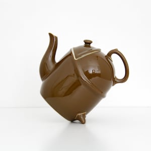 May include: A brown ceramic teapot with a distinctive, angular form. The teapot features a curved spout, a small lid, and a rounded handle. It stands on three small feet. The teapot is a solid, glossy brown colour.