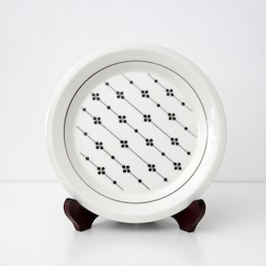 Puede incluir: Un plato de cerámica blanco con un patrón geométrico negro de líneas diagonales y pequeñas formas florales. El plato se exhibe sobre un soporte de madera marrón oscuro, sobre un fondo blanco. El diámetro del plato es de aproximadamente 20 cm.