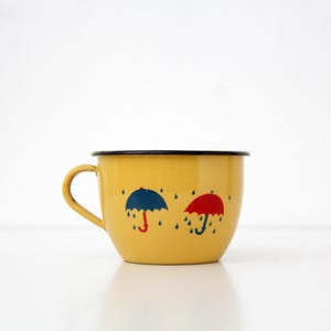 May include: A yellow enamel mug with a black rim and handle. The mug features two umbrella designs, one blue and one red, with raindrops. The mug is set against a white background.